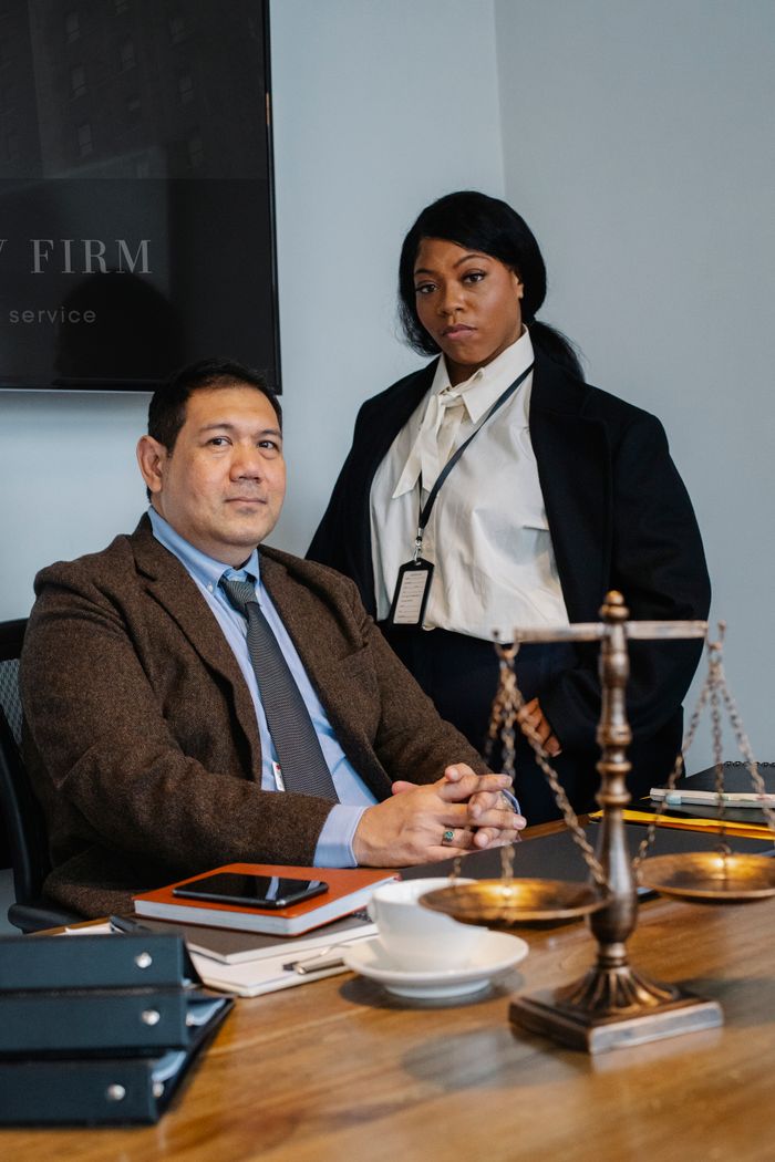 About Focused young black woman in formal clothes standing near male ethnic colleague at wooden table with documents and justice scale during work in law firm